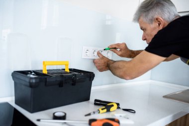 Yaşlı elektrikçi, üniformalı tamirci tornavida kullanarak mutfağa soket takıyor ya da tamir ediyor. Onarım servisi kavramı