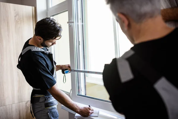 Два профессиональных работника в форме используют рулетку при измерении окна и делают заметки для установки жалюзи в помещении. Концепция строительства и обслуживания — стоковое фото