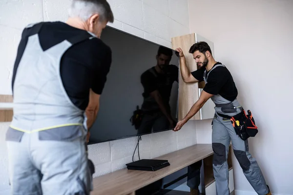 Two professional technicians, workers in uniform installing television on the wall indoors. Construction, maintenance and delivery concept — Stock Photo, Image