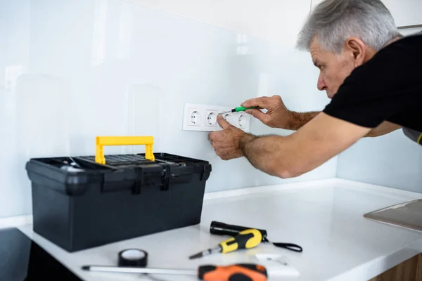 Electricista envejecido, reparador en uniforme de trabajo, instalación o fijación de enchufe en la cocina con destornillador. Servicio de reparación concepto —  Fotos de Stock