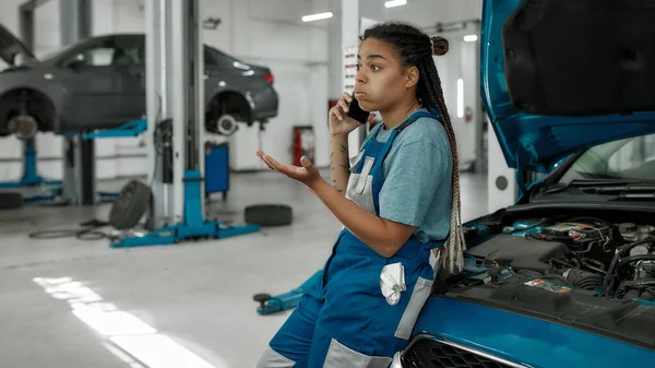 Mujer afroamericana joven, mecánico profesional hablando por teléfono, apoyado en un coche con capucha abierta en el taller de reparación de automóviles — Foto de Stock