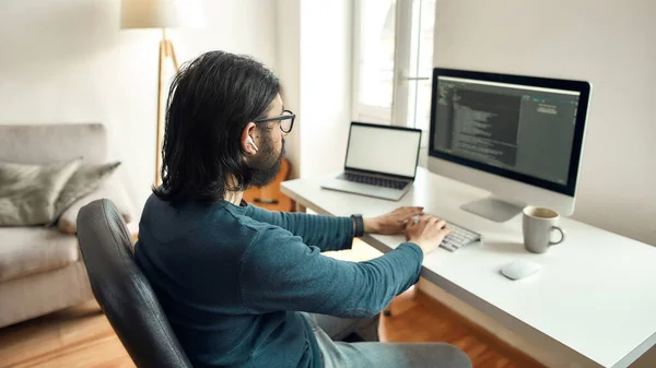 Software Programming. Side view of young male web developer wearing eyeglasses writing code on desktop computer, working from home