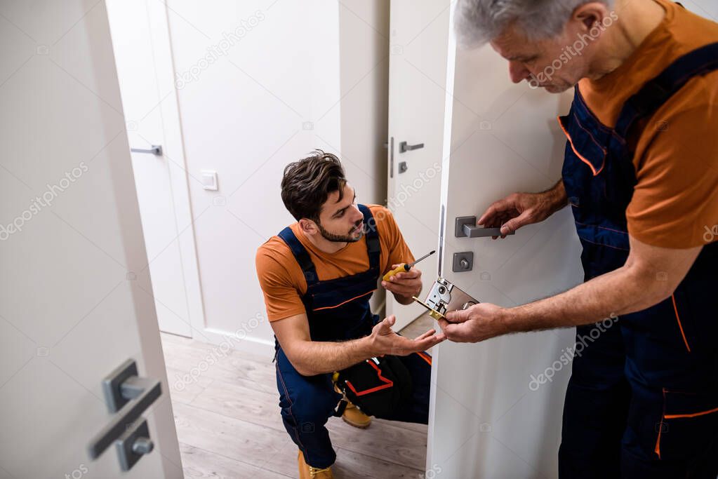 Two locksmith, repairmen, workers in uniform installing, working with house door lock using screwdriver. Repair, door lock service concept