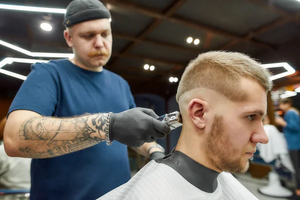 Neue trendige Frisur. Seitenansicht eines professionellen Friseurs, der mit Haarschneidemaschine arbeitet und jungen bärtigen Mann beim Friseurbesuch dient — Stockfoto