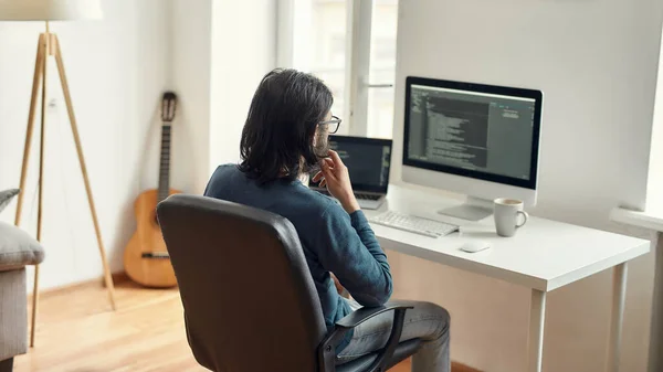Задумчивый мужчина-веб-разработчик сидит на своем рабочем месте, смотрит на экран с программным кодом, работает из дома. Использование настольного компьютера и ноутбука — стоковое фото