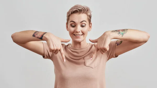 Olha para isto. Retrato de mulher tatuada com nariz perfurado e cabelo curto em camiseta bege parecendo animado apontando os dedos indicadores isolados sobre fundo claro — Fotografia de Stock