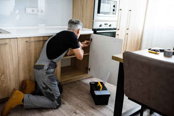 Üniformalı bir tamircinin, tornavida kullanarak mutfak dolabını tamir ederken tam boy fotoğrafı. Onarım servisi kavramı — Stok fotoğraf