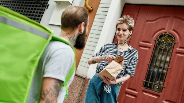 Brutal bearded delivery man with thermo backpack giving away order to a female customer, while delivering food. Courier, delivery service concept — Stock Photo, Image