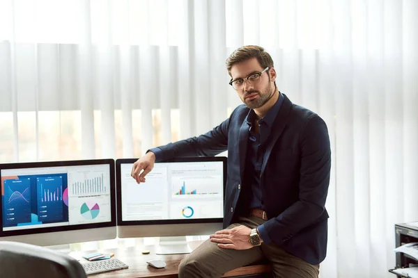 Young successful businessman in classic wear and eyeglasses leaning against a table and looking at camera, working with statistical data, graphs and charts on computer in the office — Stock Photo, Image