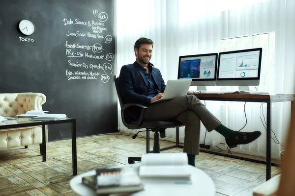 Great results. Full leghth of young happy businessman sitting at his workplace in the modern office, working on laptop and smiling — Stock Photo, Image