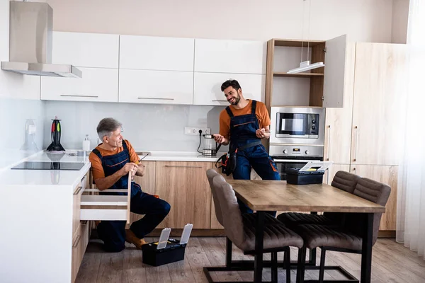 Całkowite ujęcie dwóch złotych rączki, robotnicy w mundurach rozmawiający podczas montażu szafki kuchennej za pomocą śrubokrętu w pomieszczeniu. Koncepcja naprawy i montażu mebli — Zdjęcie stockowe