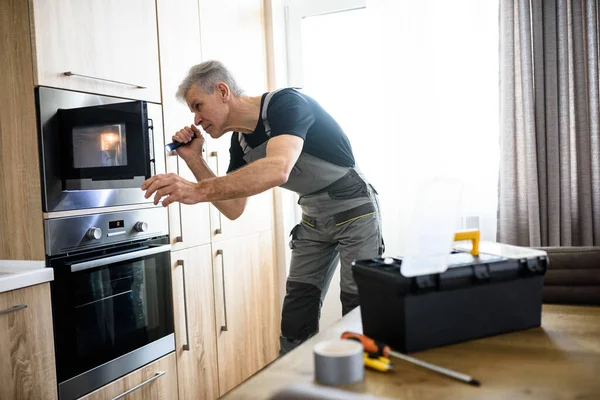 Hier zur Reparatur. Ein älterer Reparateur in Uniform arbeitet und untersucht mit Taschenlampe die kaputte Mikrowelle in der Küche. Reparaturdienstkonzept — Stockfoto