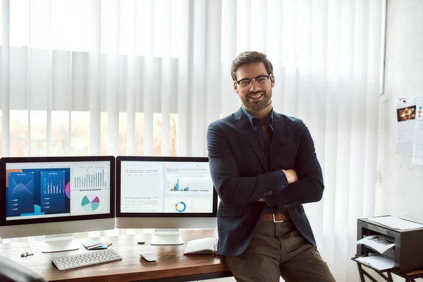 Joven analista financiero feliz en desgaste clásico y anteojos apoyados en una mesa y sonriendo a la cámara mientras trabaja con datos estadísticos —  Fotos de Stock