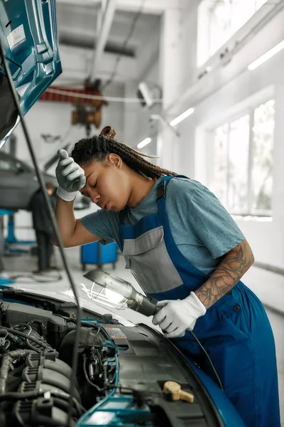 Su médico de coche. Mujer afroamericana joven, mecánico profesional limpiando la frente, examinando debajo del capó del coche con la antorcha en el taller de reparación de automóviles — Foto de Stock