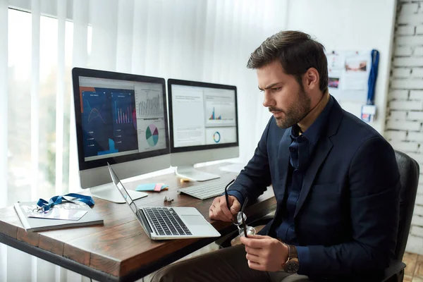 Preparing financial report. Young focused businessman or financial analyst sitting at his workplace in the modern office and working on laptop — Stock Photo, Image