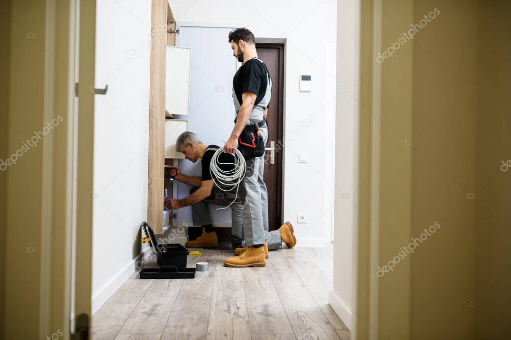 Full length shot of aged electrician, repairman in uniform working, installing ethernet cable or router in fuse box while his young colleague watching him, holding cable