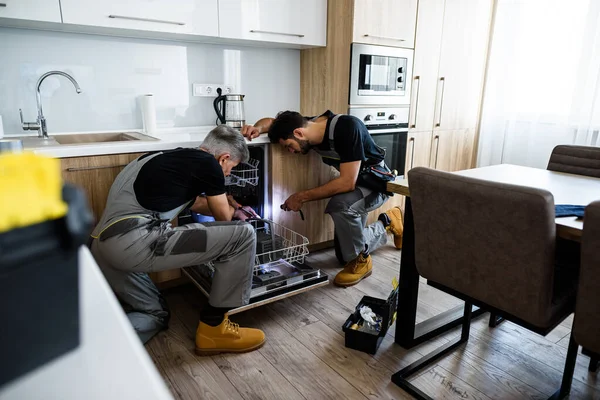 Extraordinary service. Full length shot of aged repairman fixing dishwasher in the kitchen, using screwdriver while his colleague in uniform helping him, holding flashlight. Repair service concept — Stock Photo, Image