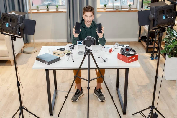 Full length shot of young male technology blogger recording video blog or vlog review of new smartphone, showing it and another one on camera at home studio. Blogging, Work from Home concept — Stock Photo, Image