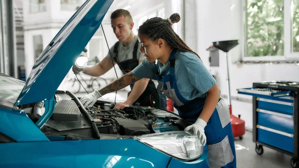 Servicios genuinos. Mujer afroamericana, mecánico profesional reparando motor de automóvil, apriete, atornille con llave mientras su colega sostiene la antorcha bajo la capucha del automóvil en la estación de servicio — Foto de Stock