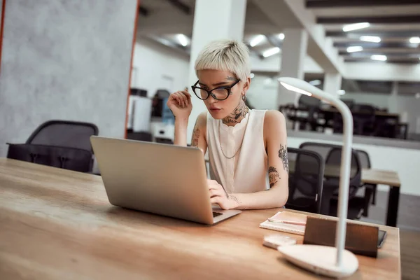 Inspiráció. Stílusos és gyönyörű tetovált üzletasszony szemüveg dolgozik laptop, miközben ül az asztalánál a modern irodában — Stock Fotó