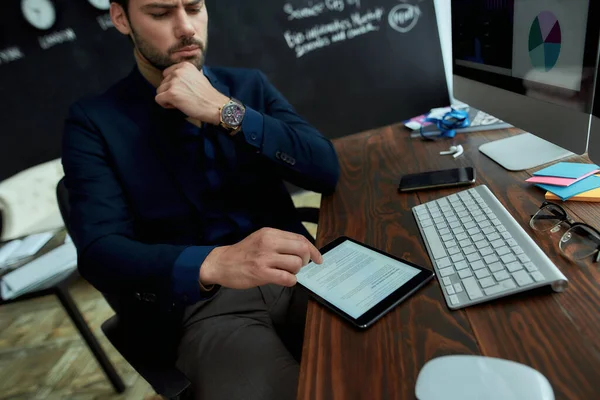 最後のニュースだ。若いビジネスマンは、現代のオフィスで彼の職場に座って、デジタルタブレット上でオンライン金融ビジネスニュースを読んで — ストック写真