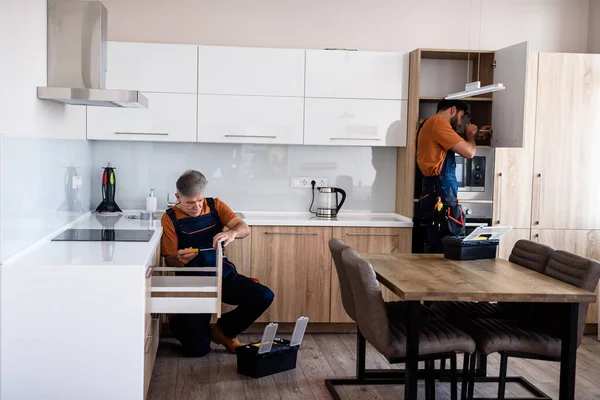 Laissez-nous travailler. Plan complet de deux bricoleurs, ouvriers en uniforme assemblant placard de cuisine, armoire à l'aide de tournevis à l'intérieur. Concept de réparation et d'assemblage de meubles — Photo