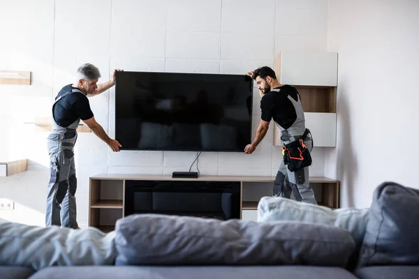 Dwóch profesjonalnych techników, pracownicy w mundurach instalujący telewizję na ścianie w pomieszczeniach. Koncepcja budowy, konserwacji i dostawy — Zdjęcie stockowe