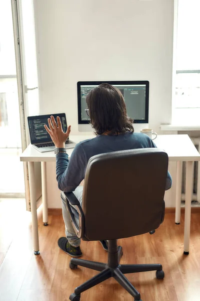 Reunión en línea con colegas. Vista posterior de desarrollador web masculino mirando webcam y saludando mientras está sentado en su lugar de trabajo y trabajando desde casa, escribir código — Foto de Stock