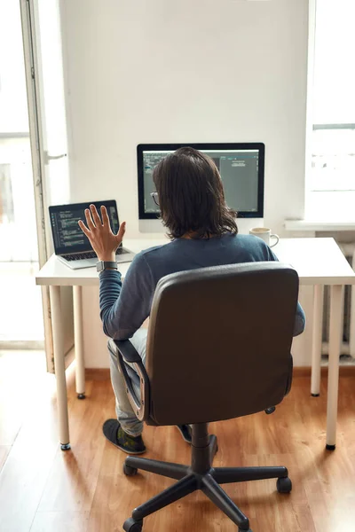 Incontro online con i colleghi. Vista posteriore di sviluppatore web maschile guardando webcam e agitando mentre seduto sul suo posto di lavoro e di lavoro da casa, scrivere codice — Foto Stock