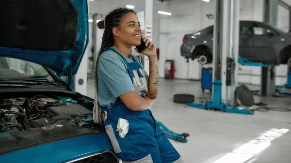 Mujer afroamericana joven, mecánico profesional hablando por teléfono, apoyado en un coche con capucha abierta en el taller de reparación de automóviles — Foto de Stock