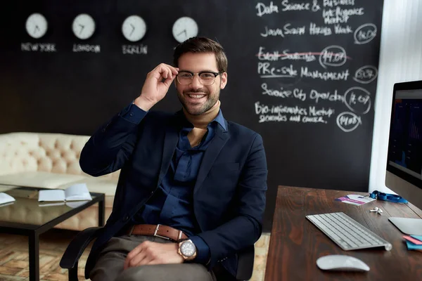 Succes. Jonge gelukkige zakenman of sales manager met bril kijkend naar de camera terwijl hij in het moderne kantoor zit met schoolbord op de achtergrond — Stockfoto