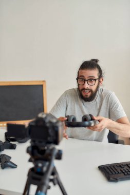 Gözlüklü erkek teknoloji blogcusu heyecanlı görünüyor, video blogu ya da video blogu kaydederken kulaklık tutuyor ve ev stüdyosunda yeni cihazlar hakkında vlog yazıyor.