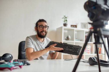 Gözlüklü erkek teknoloji blogcusu şaşırmış görünüyor, video blogu veya evdeki yeni cihazlar hakkında vlog kaydederken klavyeyi tutuyor