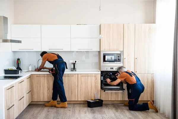 Paga y hazlo a tu manera. Dos manitas, trabajadores en uniforme de fijación, la instalación de muebles y equipos en la cocina, utilizando destornillador en el interior. concepto de reparación y montaje de muebles — Foto de Stock