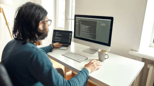 Joven desarrollador web masculino que trabaja desde casa, utilizando computadora de escritorio y computadora portátil, escribiendo código. Freelance, trabajando en línea, concepto de oficina en casa — Foto de Stock