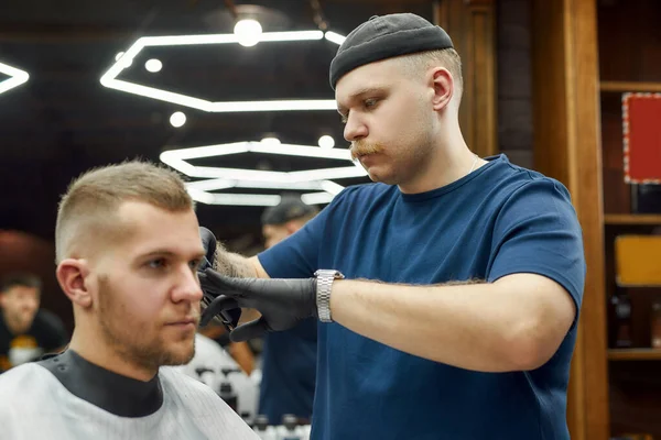 Professioneller Friseur, der im modernen Friseursalon jungen, gut aussehenden Mann dient und mit Haarschneidemaschine arbeitet. Friseur im Fokus — Stockfoto