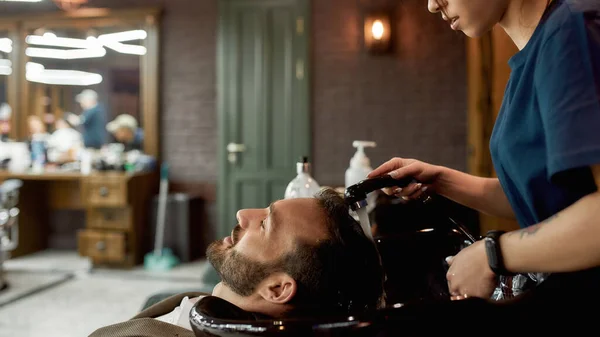 Odwiedzam salon fryzjerski. Widok z boku młodego brodatego mężczyzny relaksującego się podczas gdy kobieta fryzjer myje włosy w nowoczesnym salonie fryzjerskim — Zdjęcie stockowe