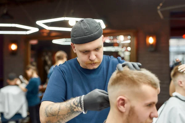 Saç makasıyla çalışan profesyonel berber, genç adam için modaya uygun saç kesimi yapıyor. Dövmeli bir adama odaklan. — Stok fotoğraf