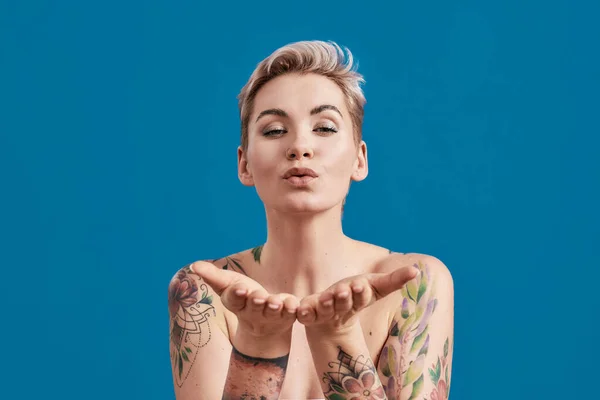 Retrato de una joven atractiva mujer semidesnuda tatuada con la piel perfecta soplando beso a la cámara aislada sobre fondo azul — Foto de Stock