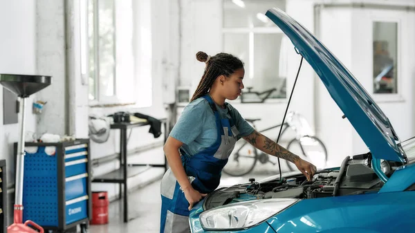 Calidad en cada trabajo. Mujer afroamericana joven, mecánico profesional mirando, examinando debajo de la capucha del coche en el taller de reparación de automóviles — Foto de Stock