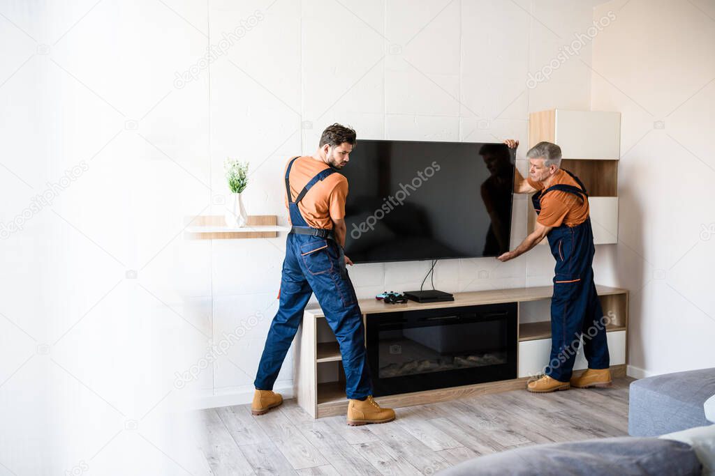 Delivered on time. Two handymen, workers in uniform hanging, installing tv television on the wall indoors. Repair and assembly service concept