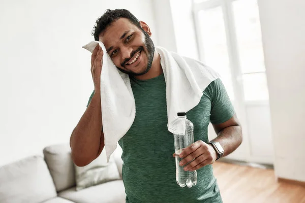 Κουρασμένος αλλά ευτυχισμένος. Νεαρός άνδρας χαμογελώντας στην κάμερα, κρατώντας το μπουκάλι του νερού και σκουπίζοντας τον ιδρώτα με πετσέτα ενώ ξεκουράζεται μετά την προπόνηση στο σπίτι. Αθλητισμός, υγιεινός τρόπος ζωής — Φωτογραφία Αρχείου