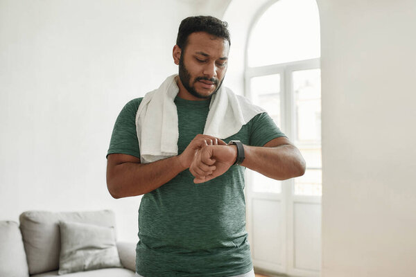 Check the result. Young active man with white towel around neck checking time or results in smartwatch after doing morning workout at home