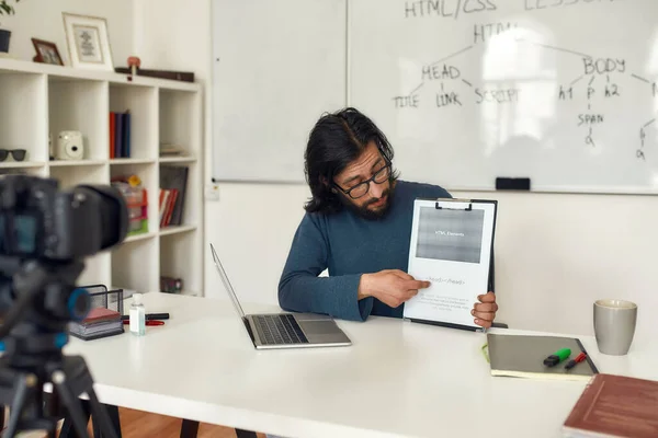 Νεαρός καυκάσιος δάσκαλος φορώντας γυαλιά που διδάσκει πώς να κωδικοποιεί HTML, να κάθεται στο χώρο εργασίας του και να δίνει online μάθημα. Εγγραφή blog βίντεο — Φωτογραφία Αρχείου