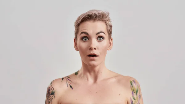 Choque. Retrato de una joven atractiva mujer tatuada con la nariz perforada y el pelo corto mirando sorprendida o sorprendida ante la cámara aislada sobre fondo claro — Foto de Stock