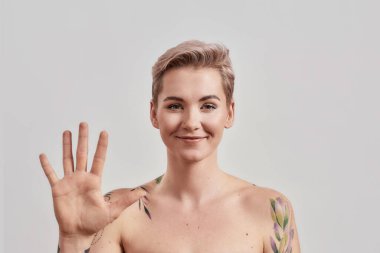 Selamlar. Neşeli, yarı çıplak, burnu delinmiş, kısa saçlı, arka planda izole bir şekilde kameraya el sallayan kadın portresi.