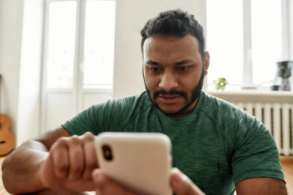 Κοντινό πλάνο του νεαρού άνδρα που χρησιμοποιεί την εφαρμογή smartphone ενώ κάνει πρωινή προπόνηση στο σπίτι. Ο πρωτοετής χαλαρώνει μετά την προπόνηση. Online προσωπικός γυμναστής στο κινητό τηλέφωνο — Φωτογραφία Αρχείου