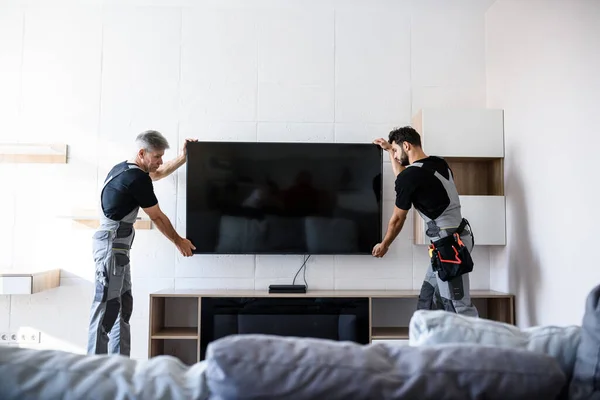 2人のプロの技術者は、壁に屋内でテレビを設置する制服の労働者。建設、メンテナンス、配送のコンセプト — ストック写真