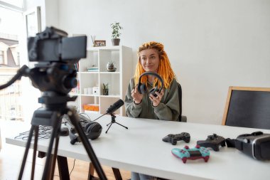 Rastalı kadın teknoloji blogcusu kameraya bakıyor, kulaklık tutuyor ve evdeki yeni cihazların video incelemelerini kaydediyor.