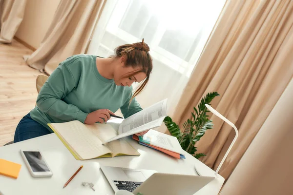 Молодая женщина дистанционно учится. Молодая студентка сидит за столом и читает пособие, использует ноутбук, делает заметки во время учебы дома — стоковое фото